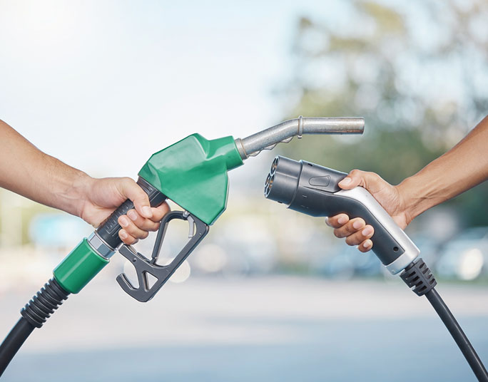 Two hands holding fuel nozzles facing each other, one green gasoline pump nozzle and one black electric vehicle charging connector.