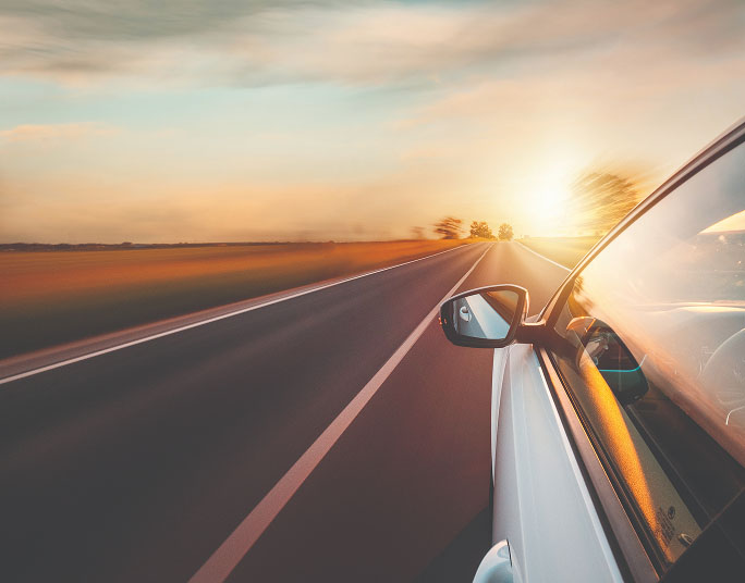 A white car driving on a two-lane road during sunset and surrounding landscape