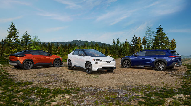 Three Subaru electric SUVs parked on a grassy clearing with a forest and hills in the background under a partly cloudy sky.
