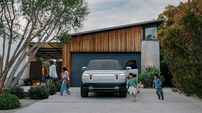 A modern house with wooden and concrete exterior featuring a silver Rivian R2 parked in driveway.