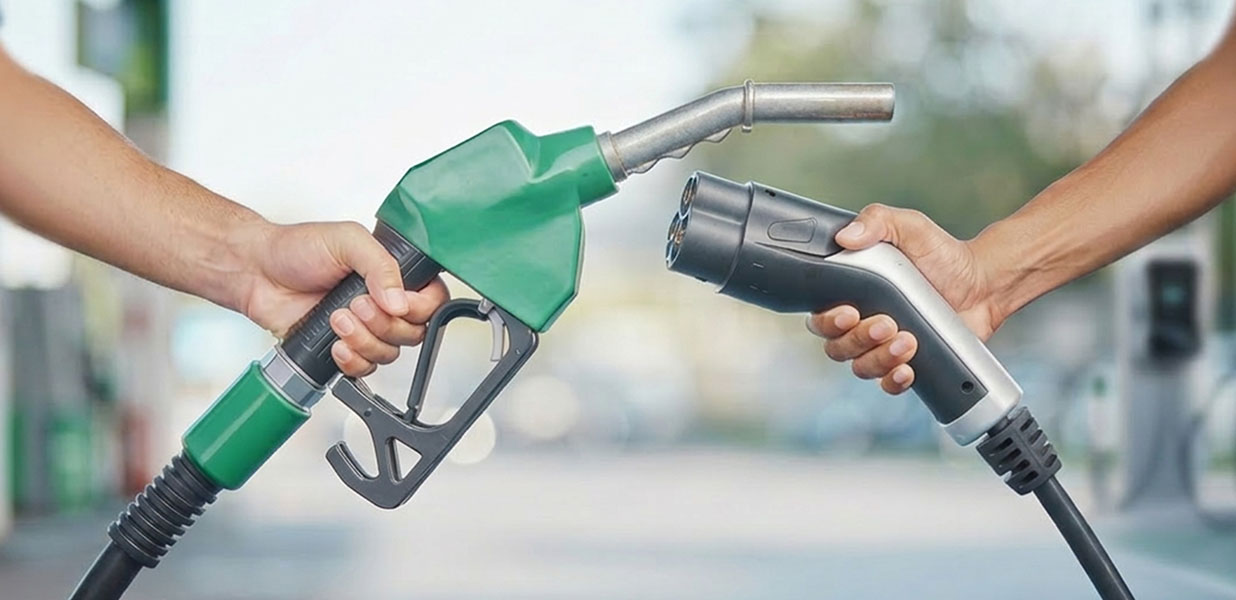Two hands holding fuel nozzles facing each other, one green gasoline pump nozzle and one black electric vehicle charging connector.