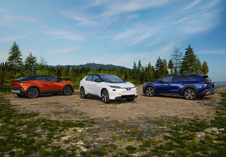 Three Subaru electric SUVs parked on a grassy clearing with a forest and hills in the background under a partly cloudy sky.

