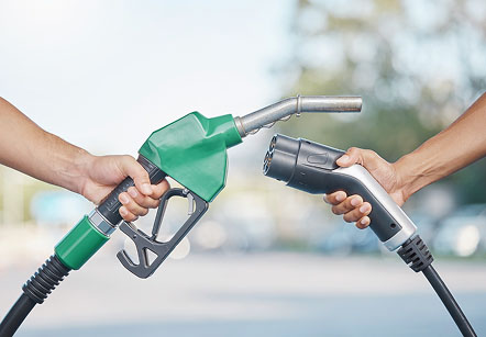 Two hands holding fuel nozzles facing each other, one green gasoline pump nozzle and one black electric vehicle charging connector.