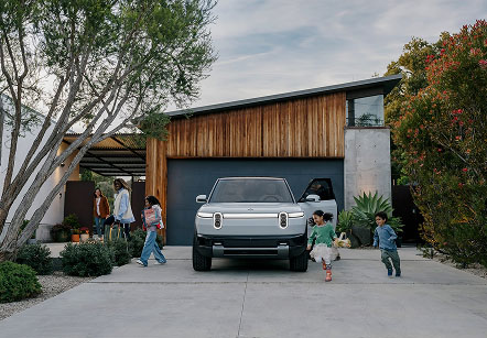 A modern house with wooden and concrete exterior featuring a silver Rivian R2 parked in driveway.