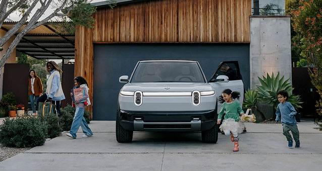 A modern house with wooden and concrete exterior featuring a silver Rivian R2 parked in driveway.