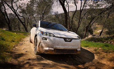 A white 2026 Subaru Solterra electric SUV driving on a dirt trail surrounded by trees and hills.