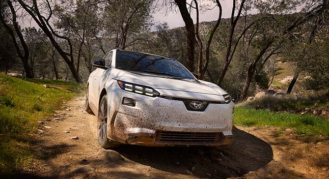 A white 2026 Subaru Solterra electric SUV driving on a dirt trail surrounded by trees and hills.