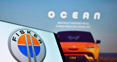 A tablet displaying the Fisker electric vehicle logo is held in front of a car