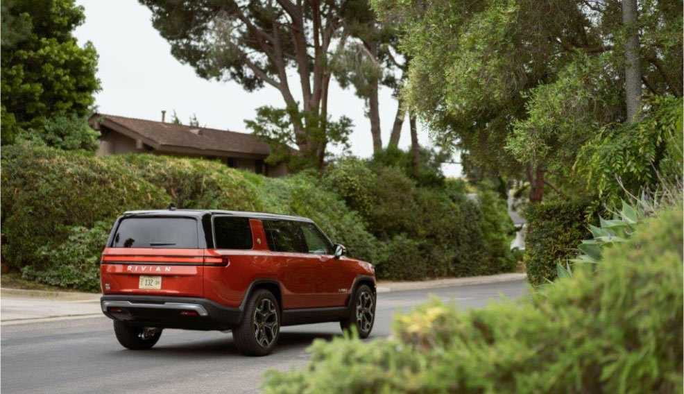 A Rivian electric vehicle driving down a street with a lot of foliage