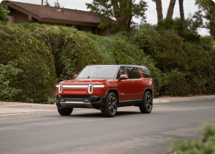 A Rivian R1S electric vehicle driving down a street lined by tall bushes