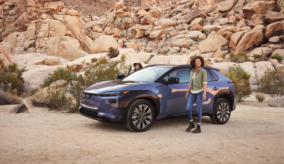 A woman standing beside a blue Subaru sedan in a rocky desert landscape