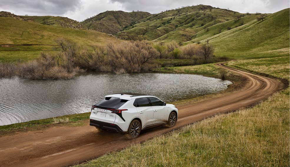 A white Subaru driving along a curved dirt road beside a pond in rolling green hills
