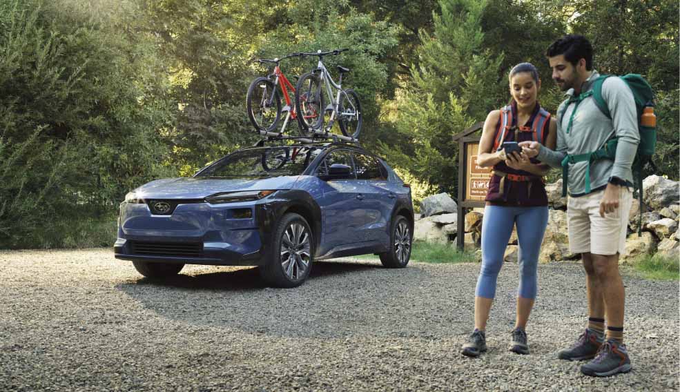 A couple preparing beside a blue Subaru parked on a gravel trail, with two bicycles mounted on its roof