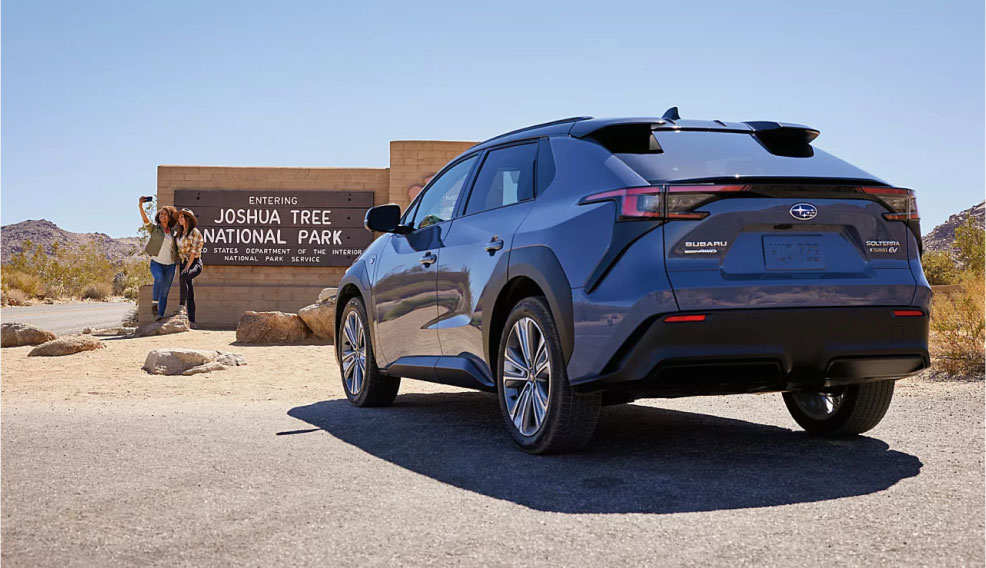 A Subaru electric vehicle parked in front of the entrance sign to Joshua Tree National Park, where two people are taking a selfie in front